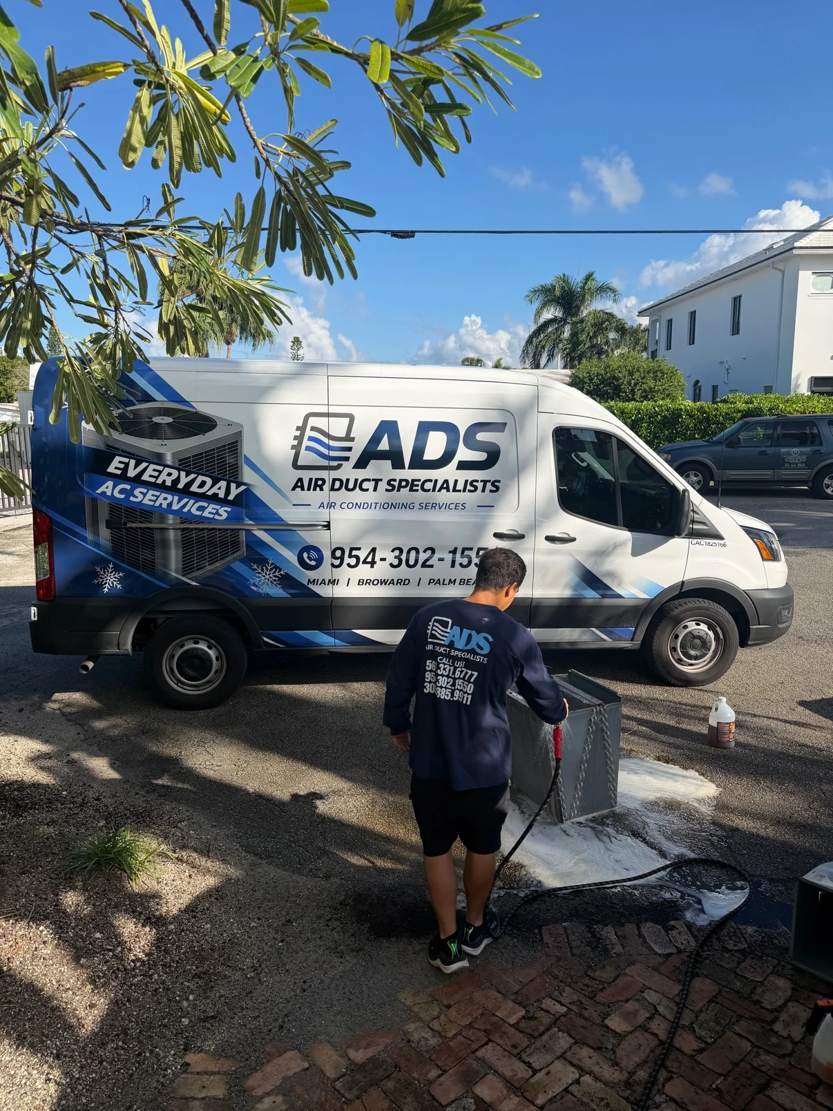 Technician hosing down a removed evaporator coil beside an Air Duct Specialists van, foam runoff on the driveway — Air Duct Specialists LLC