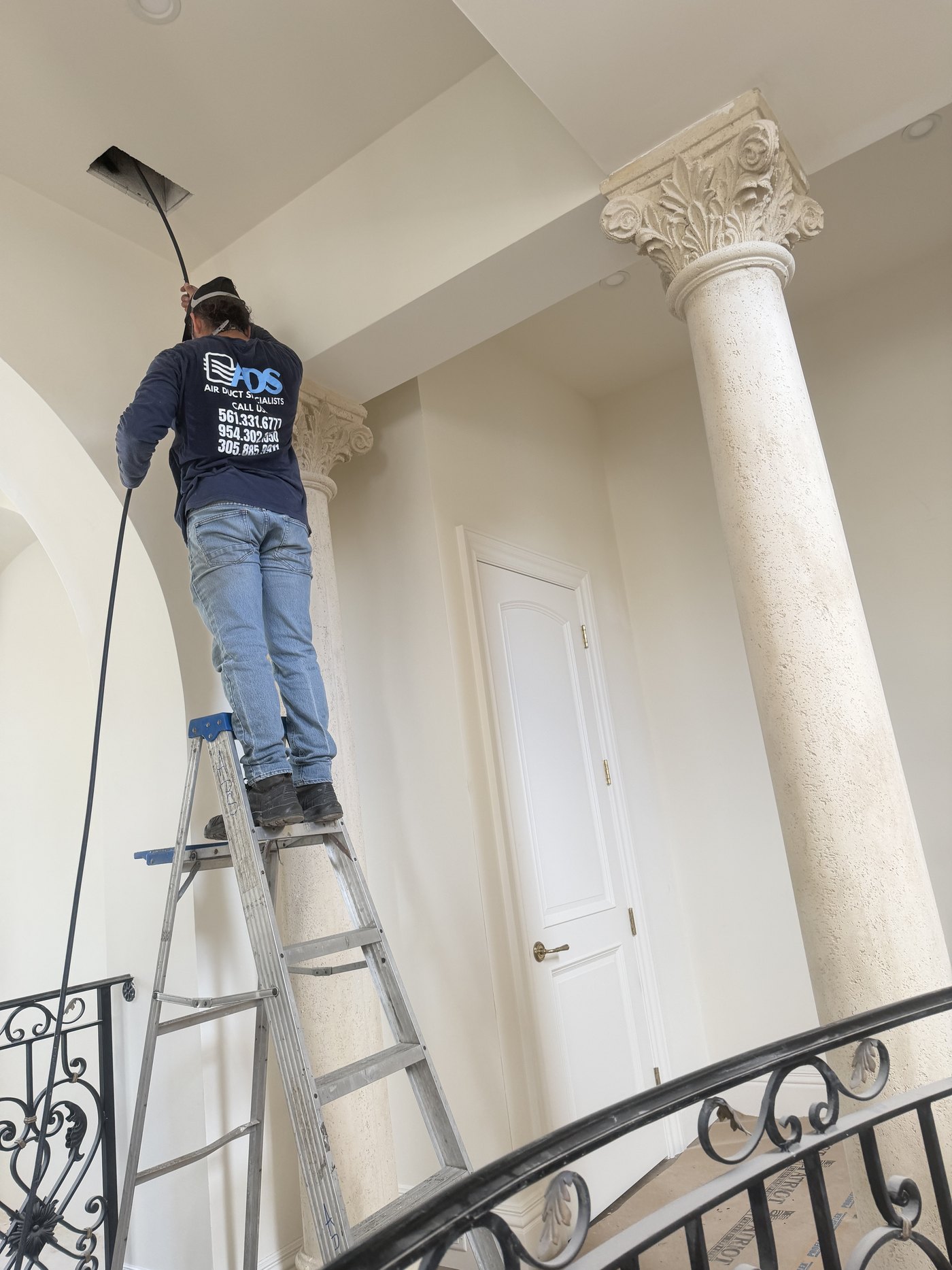 Technician reaching a high ceiling supply register with collection hose attached during estate home duct cleaning in Boca Raton