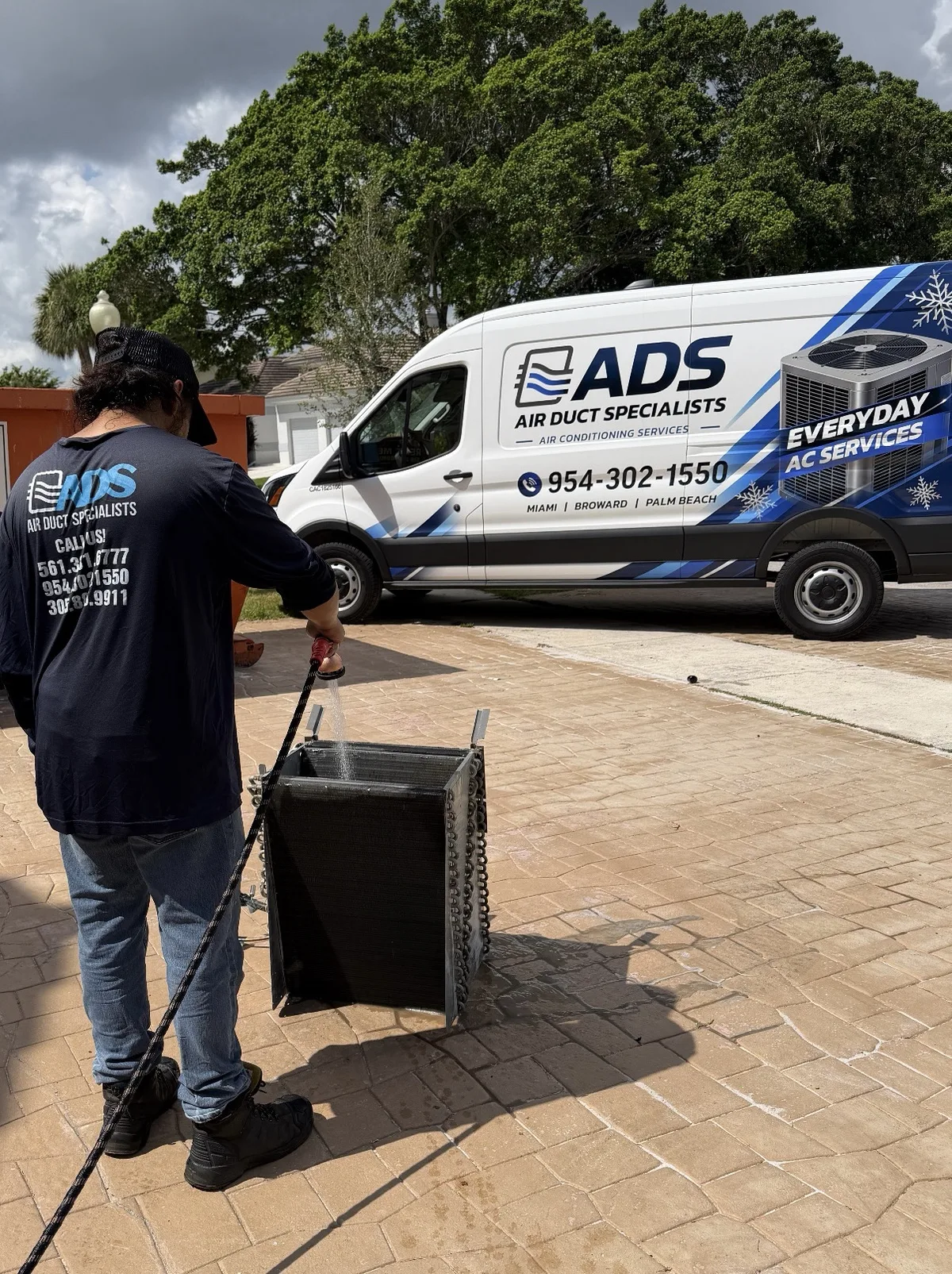 Air Duct Specialists technician hosing a removed evaporator coil on a driveway with the company van behind
